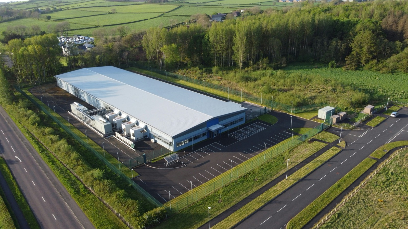 Aerial view of a modern data centre with rooftop cooling units, set in green countryside beside a road.