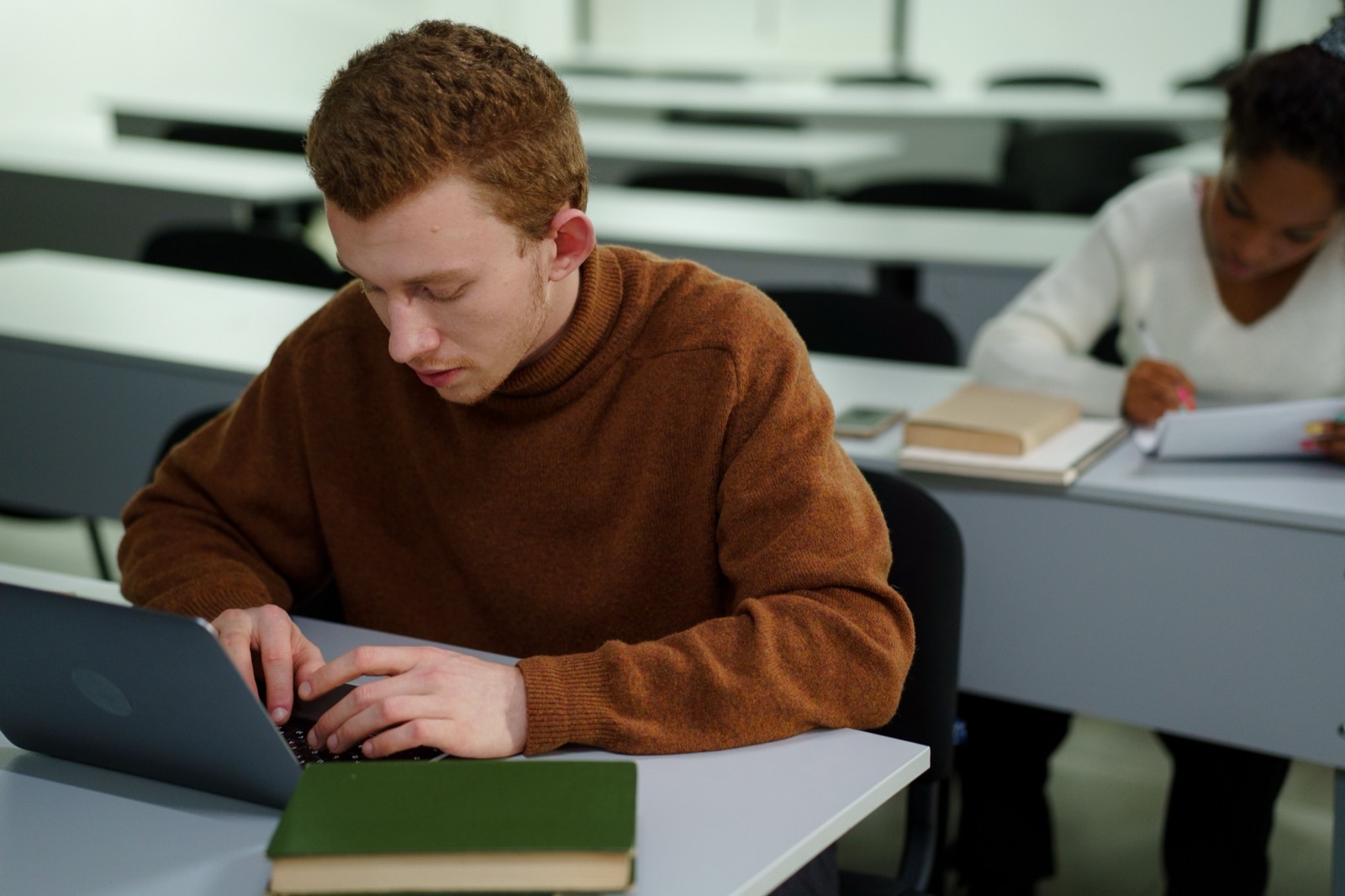 A student typing on a laptop at a lecture-hall desk, a stack of books beside them and another student working behind.
